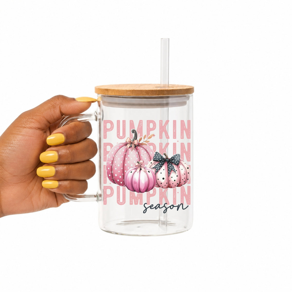 A hand with yellow nail polish holds a clear 17oz glass handled mug with a bamboo lid and clear straw. The tumbler features a pink and white 'Pumpkin Season' graphic with watercolor pumpkins.