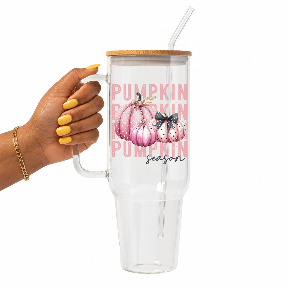 A hand with yellow nail polish holds a clear glass 40oz tumbler with a bamboo lid and straw. The tumbler features a pink 'Pumpkin Season' graphic with watercolor pumpkins.