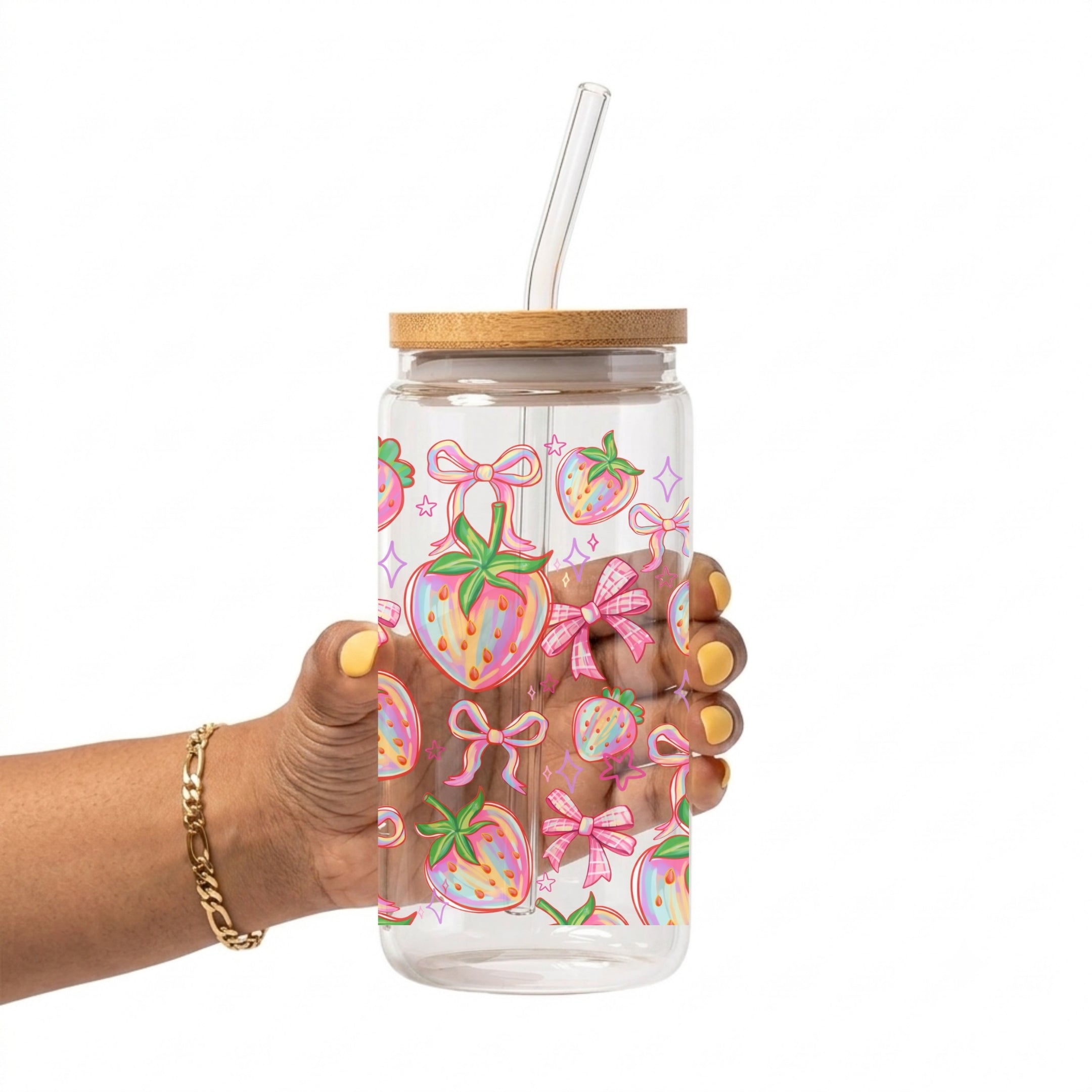A hand holding a clear glass cup with a bamboo lid and clear straw. The cup is decorated with a colorful pattern of pink bows and strawberries.
