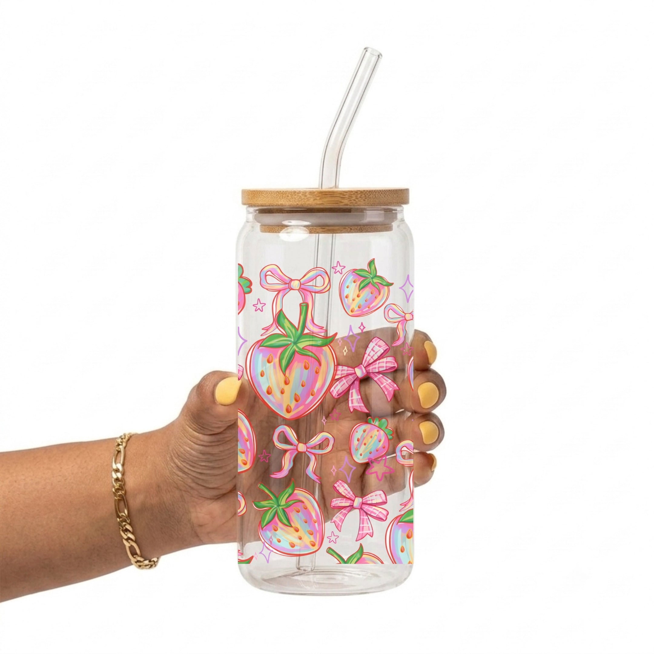 A hand holding a clear 20oz glass cup with a bamboo lid and clear straw, decorated with a colorful pattern of pink strawberries and bows, against a white background.
