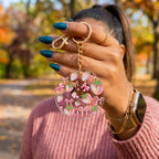 A person outdoors holds up a round clear acrylic keychain charm decorated with pink bows, pastel strawberries, and green leaves. The keychain has a gold-tone metal clasp and chain.