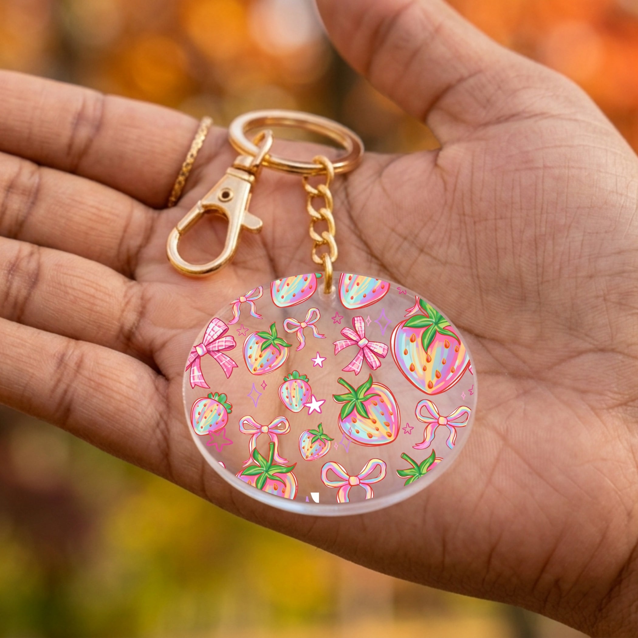 A round acrylic keychain with a gold clasp and chain, decorated with colorful strawberry and bow patterns, held in a person's hand against a blurred autumn background.