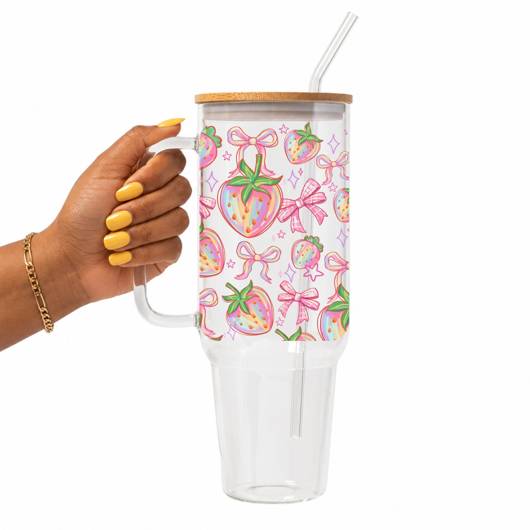 A hand with yellow nail polish holds a clear handled 40oz glass tumbler with a bamboo lid and clear straw. The tumbler features a white background decorated with a colorful pattern of pink bows and strawberries.