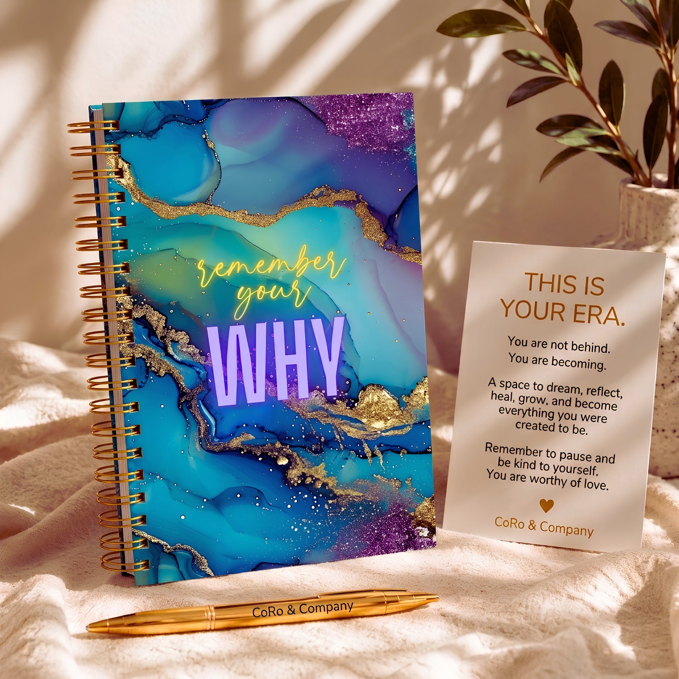 A spiral-bound self-love journal with a blue, purple, and teal marbled cover and gold foil accents sits on a soft cream fabric beside a printed affirmation card and a gold pen. The notebook cover reads 'REMEMBER YOUR WHY', and the pen and card show the brand CoRo & Company.
