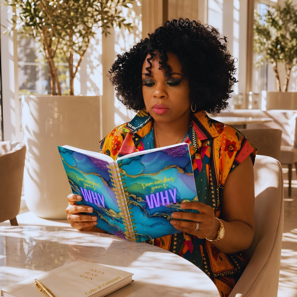Nik Austin with curly black hair sits in a bright, sunlit room reading a colorful journal titled 'REMEMBER YOUR WHY'. She wears a vibrant multicolor patterned dress and gold jewelry, seated in a light beige chair at a table with another book on it.