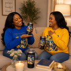 Two SGRho women sitting on a beige couch in a cozy living room setting, each holding a black insulated travel tumbler with a blue and yellow Greek letter design. They are wearing matching blue and yellow sweatshirts with the same graphic.