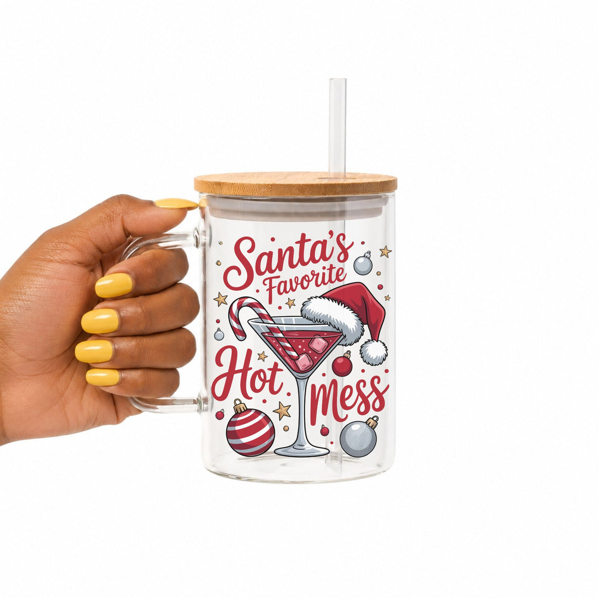 A hand with yellow nail polish holds a clear glass mug with a bamboo lid and straw. The mug features a festive Christmas graphic with the text 'Santa's Favorite Hot Mess' in red and white, alongside illustrations of a martini glass, candy cane, ornaments, and a Santa hat.