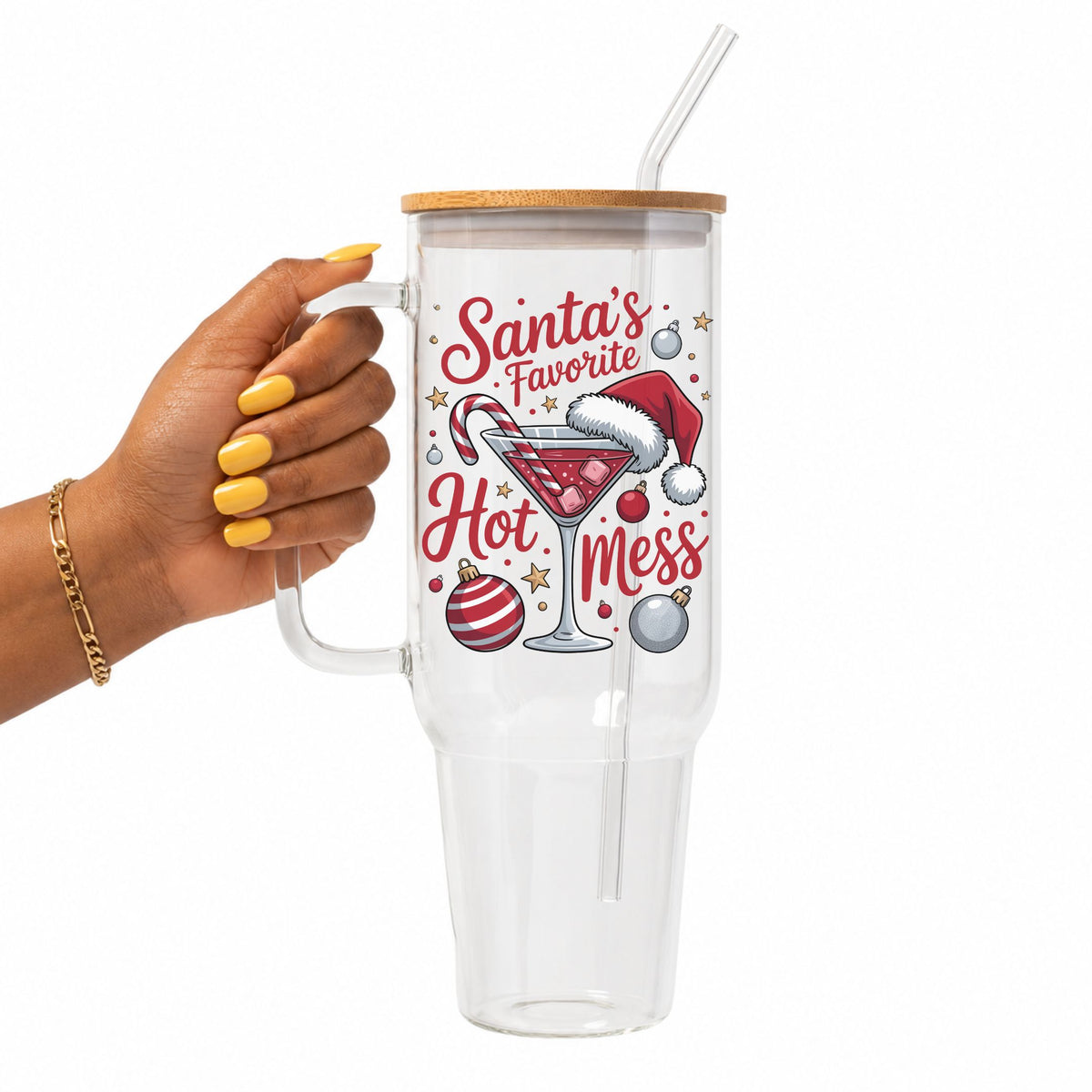 A hand with yellow nails holds a clear handled tumbler with a bamboo lid and straw. The tumbler features a festive graphic with the text 'Santa's Favorite Hot Mess' and holiday illustrations including a martini glass, candy cane, Santa hat, ornaments, and stars.
