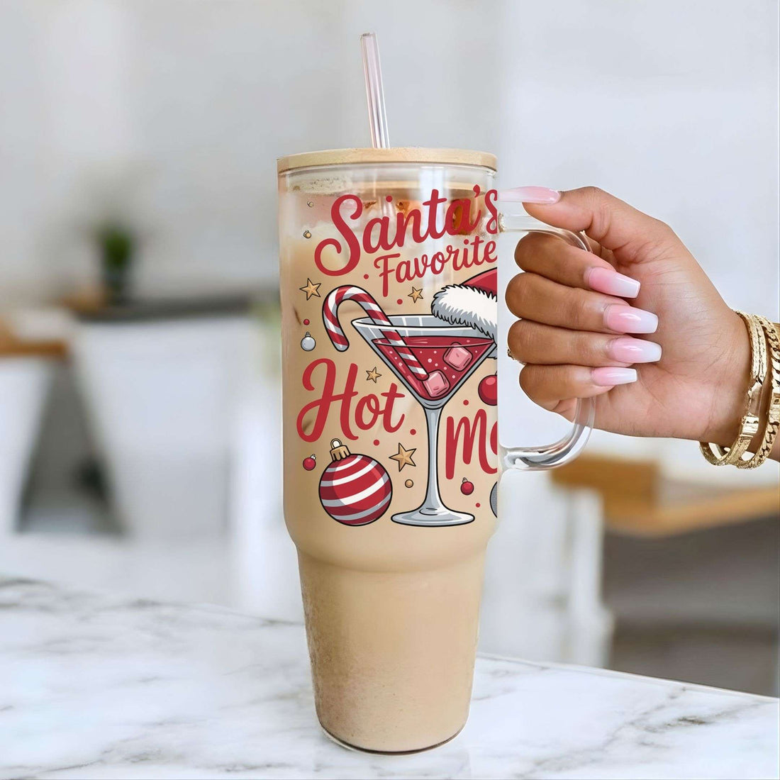 A person's hand with pink nails holds a clear handled tumbler with a straw, filled with a light brown iced drink. The tumbler has a festive red and gold holiday graphic that reads 'Santa's Favorite Hot Mocha' with illustrations of a martini glass, candy cane, stars, and ornaments.