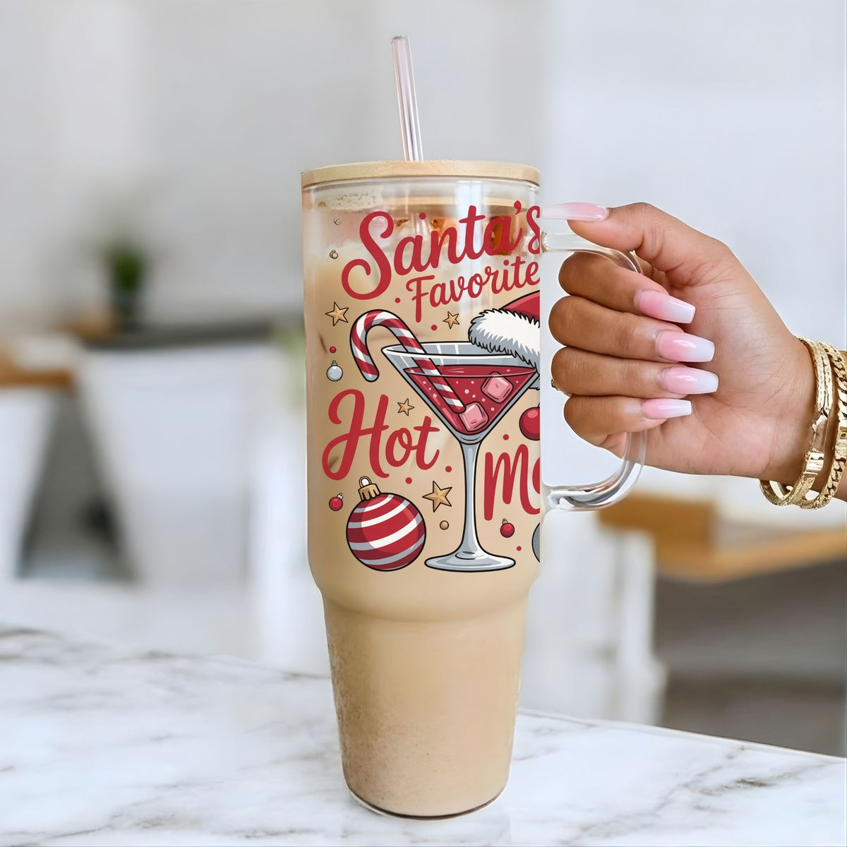 A person's hand with pink nails holds a clear handled tumbler with a straw, filled with a light brown iced drink. The tumbler has a festive red and gold holiday graphic that reads 'Santa's Favorite Hot Mocha' with illustrations of a martini glass, candy cane, stars, and ornaments.