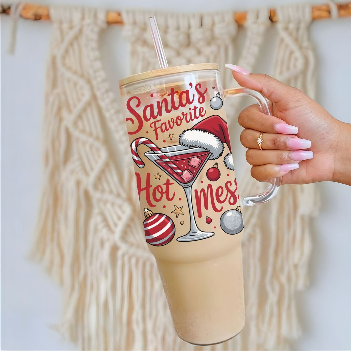 A hand with pink manicured nails holds a large insulated tumbler with a handle and straw. The tumbler has a festive Christmas design featuring the text 'Santa's Favorite Hot Mess' with illustrations of a martini glass, candy cane, ornaments, and a Santa hat.