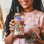 A woman holding a clear glass tumbler with a bamboo lid and glass straw, decorated with colorful self-affirmation quotes. She is wearing a pink bow-patterned t-shirt and a leopard-print scrunchie.