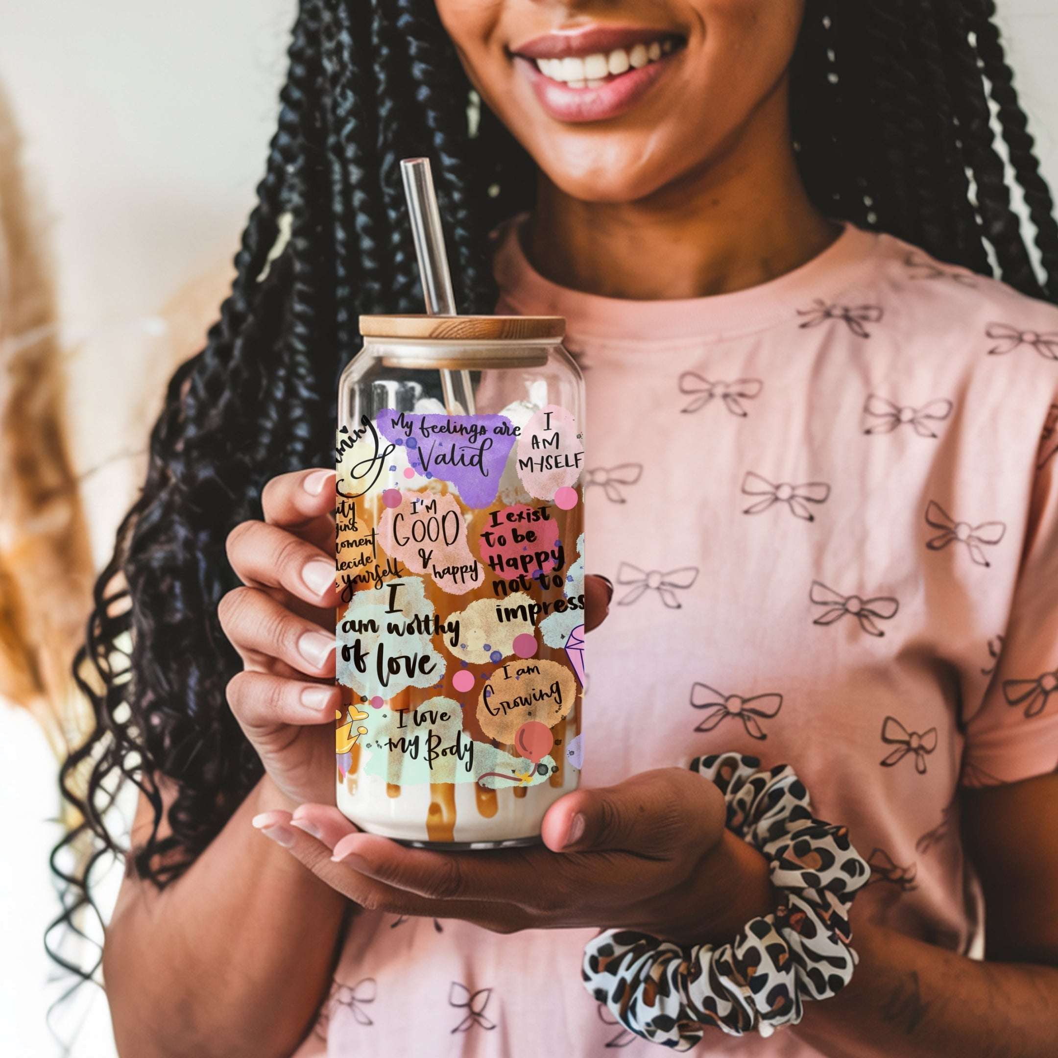A woman holding a clear glass tumbler with a bamboo lid and glass straw, decorated with colorful self-affirmation quotes. She is wearing a pink bow-patterned t-shirt and a leopard-print scrunchie.