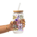A hand holding a clear glass tumbler with a bamboo lid and clear straw. The tumbler is decorated with various colorful illustrations and motivational quotes including 'I am enough', 'I trust the process', and 'I am loved'.