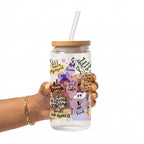 A hand holding a clear glass tumbler with a bamboo lid and clear straw. The tumbler is decorated with various colorful illustrations and motivational quotes including 'I am enough', 'I trust the process', and 'I am loved'.