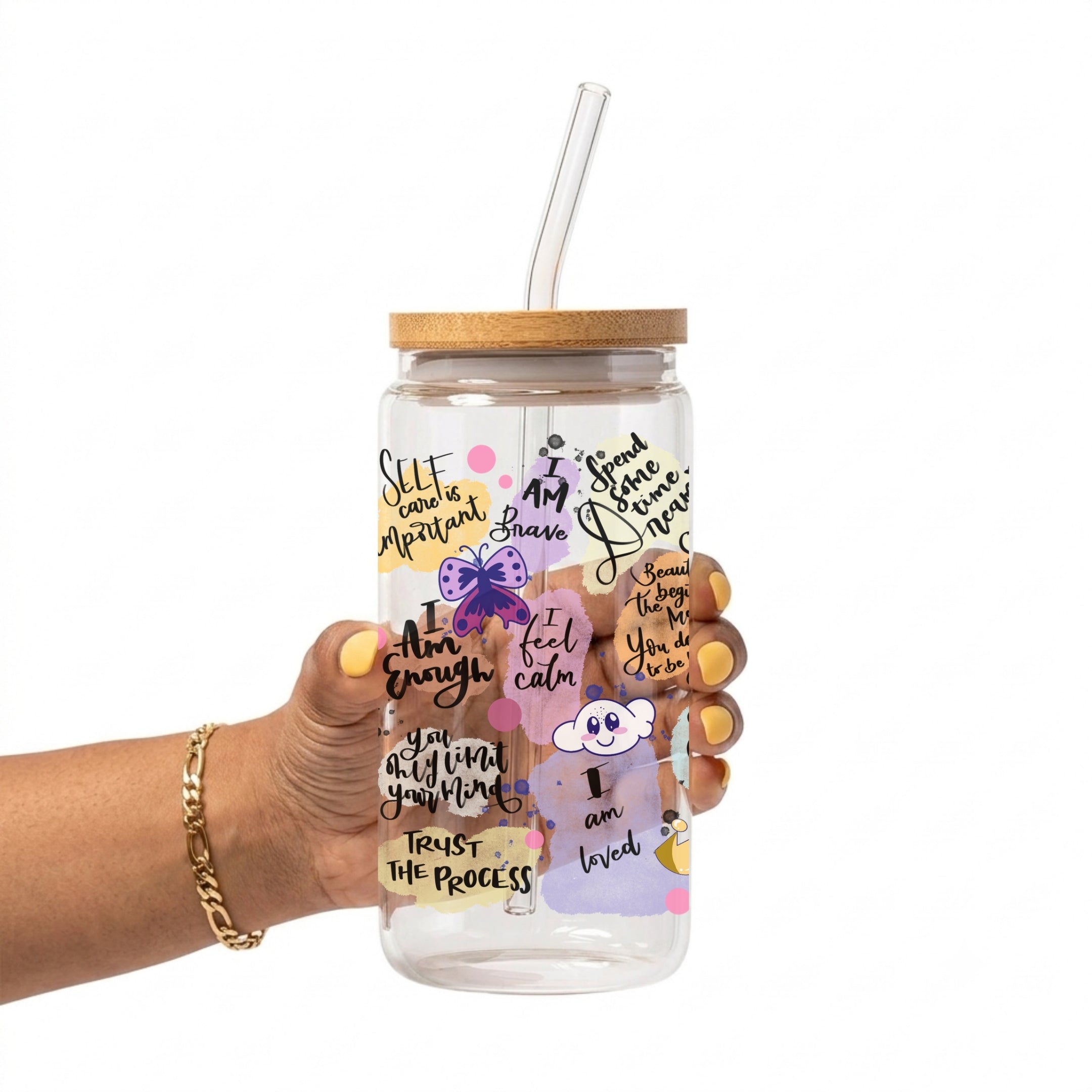 A hand holding a clear glass tumbler with a bamboo lid and clear straw. The tumbler is decorated with various colorful illustrations and motivational quotes including 'I am enough', 'I trust the process', and 'I am loved'.