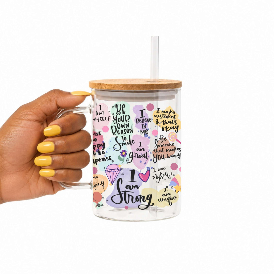 A hand with yellow nail polish holds a clear glass mug with a light wood lid and clear straw. The mug is decorated with colorful text, flowers, and icons, including the phrase 'I am Strong'.