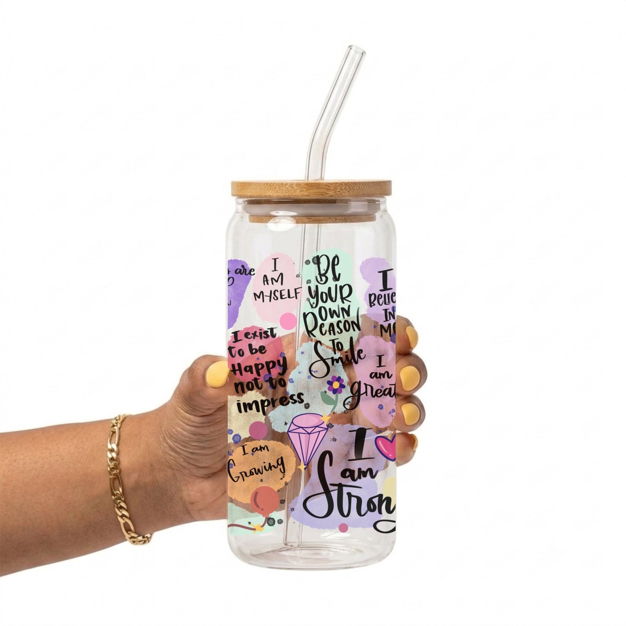 A hand holding a clear glass tumbler with a bamboo lid and clear straw. The tumbler is decorated with colorful pastel graphics and motivational text including 'I am Strong', 'Be your own person', and 'Smile when others are sad'.