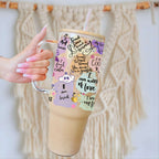 A hand with pink manicured nails holds a large insulated tumbler with a clear glass handle and straw. The tumbler has a light wood-look lid and base, decorated with a pastel collage of quotes, doodles, and small cartoon characters.