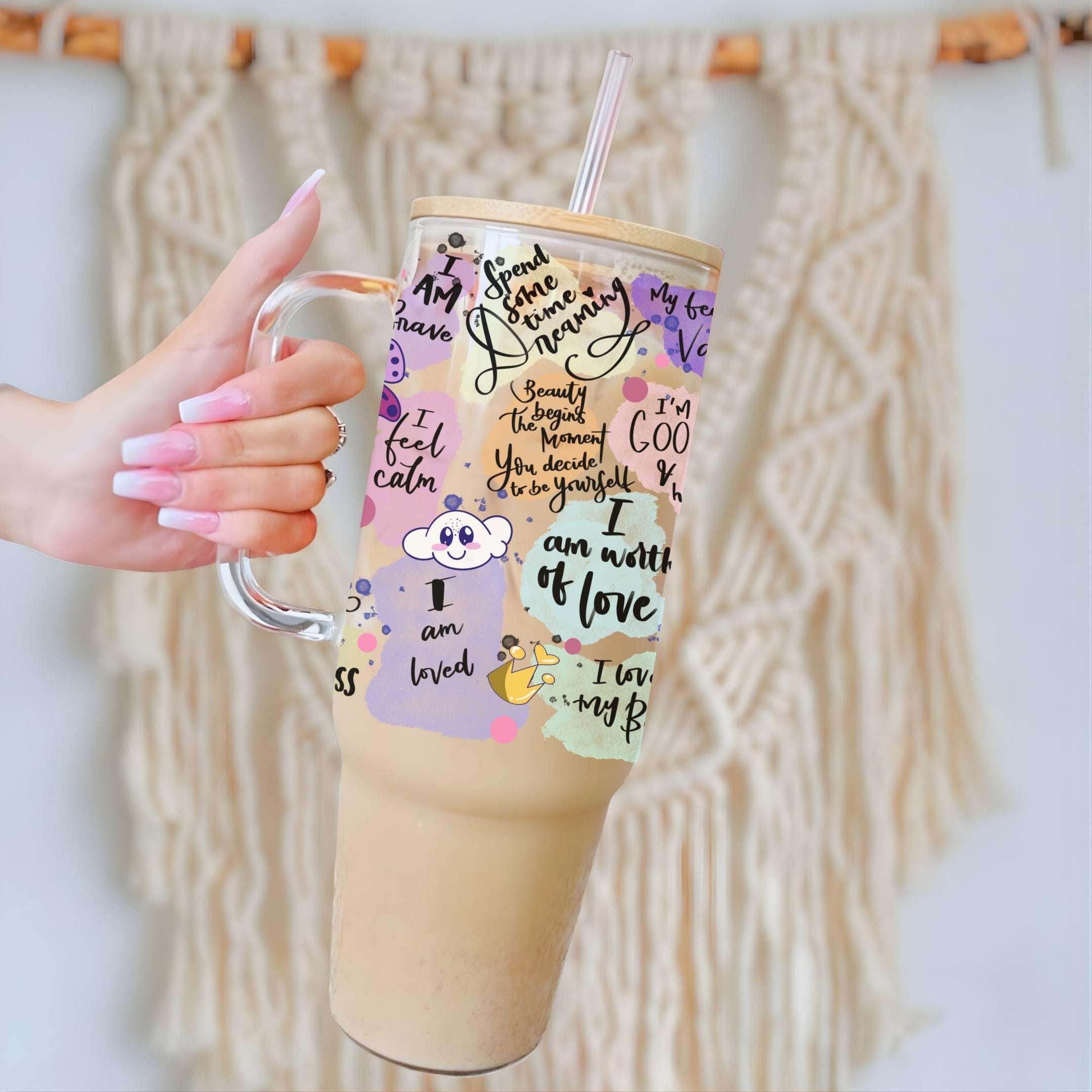 A hand with pink manicured nails holds a large insulated tumbler with a clear glass handle and straw. The tumbler has a light wood-look lid and base, decorated with a pastel collage of quotes, doodles, and small cartoon characters.