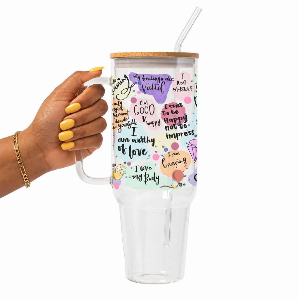 A hand with yellow nail polish holds a clear handled tumbler with a bamboo lid and clear straw. The tumbler is decorated with colorful pastel watercolor shapes and positive self-affirmations like 'My feelings are Valid' and 'I am worthy of love'.