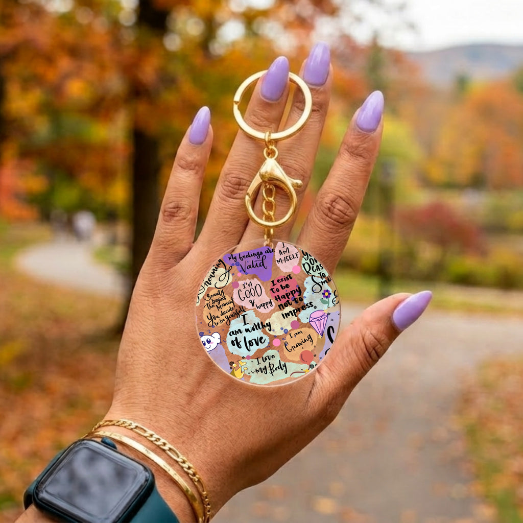 A hand with lavender nail polish holds a round acrylic keychain featuring colorful motivational quotes and illustrations. The keychain has a gold-tone split ring and lobster clasp, set against a blurred autumn park background.