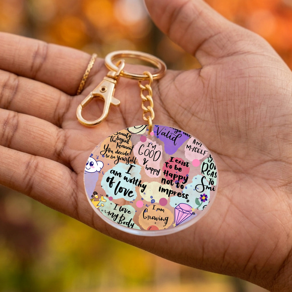 A round acrylic keychain held in a person's hand, featuring a colorful collage of motivational quotes, doodles, and small illustrations on a gold-tone clasp and chain.