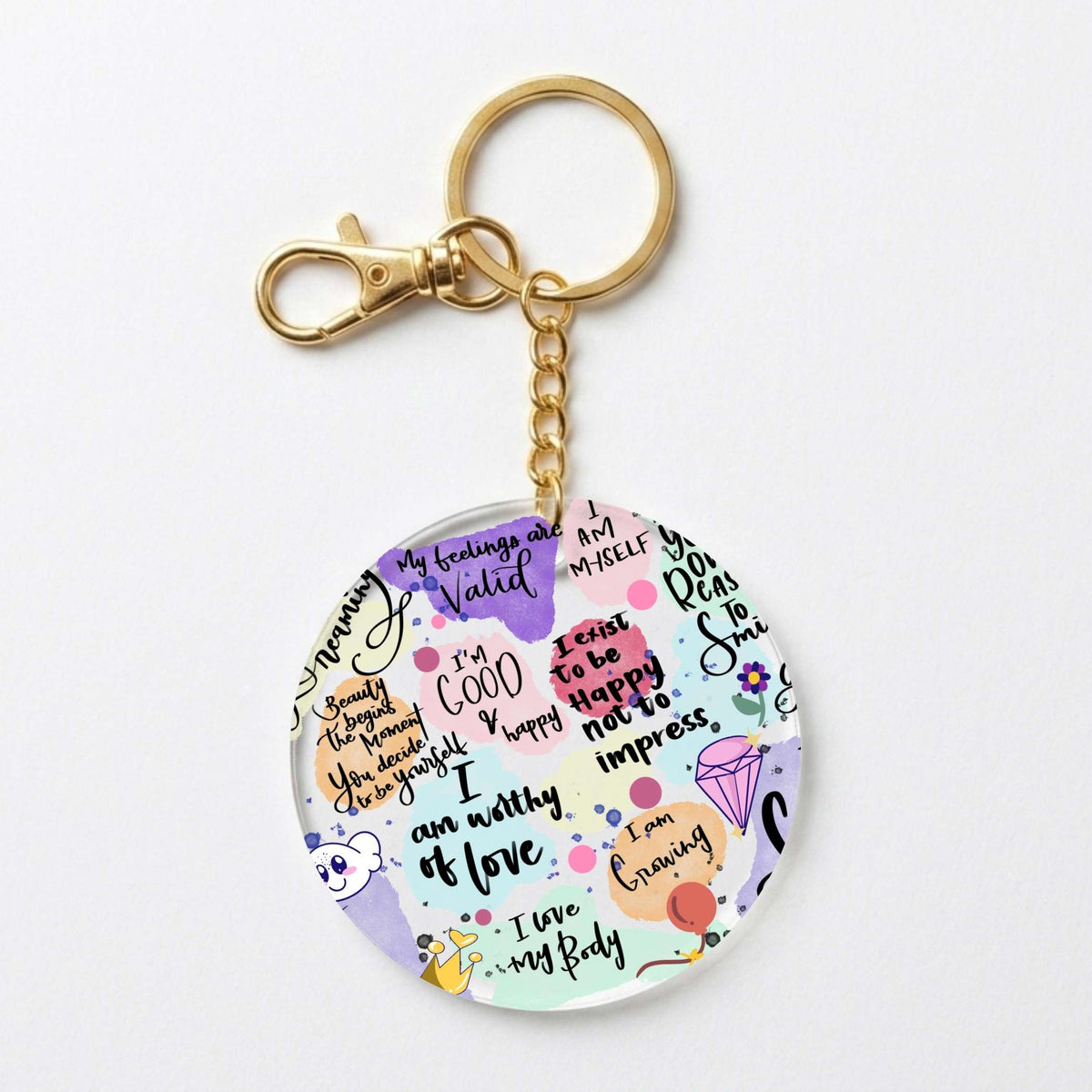 A round acrylic keychain charm featuring colorful self-affirmation quotes, doodles, and small illustrations, attached to a gold-tone split ring and clasp on a white background.