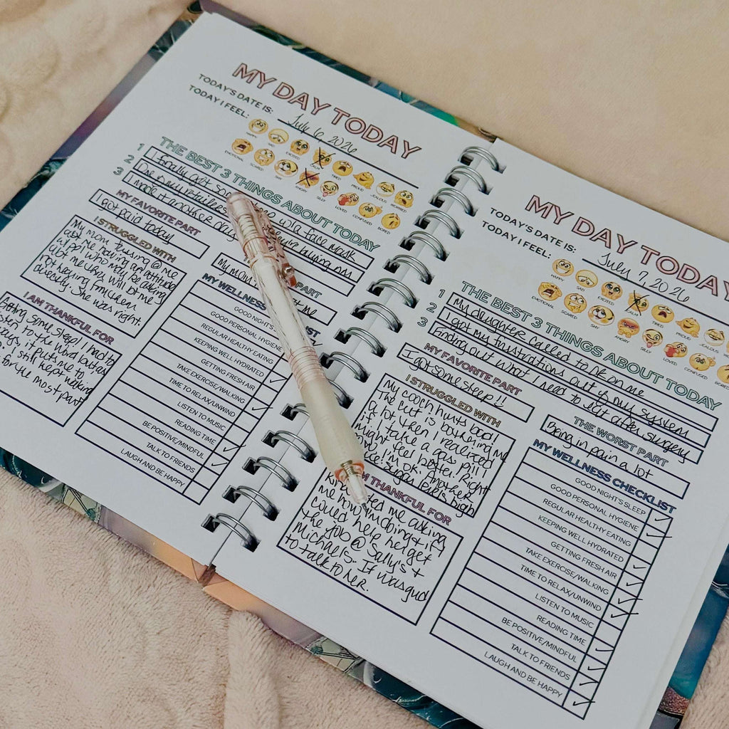 A spiral-bound self-love journal page spread open on a soft beige fabric surface, with a clear pen resting across the pages. The planner has printed sections, checklist boxes, and emoji icons, and the pages are slightly angled in the frame.