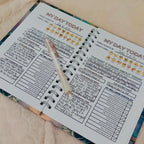 A spiral-bound self-love journal page spread open on a soft beige fabric surface, with a clear pen resting across the pages. The planner has printed sections, checklist boxes, and emoji icons, and the pages are slightly angled in the frame.