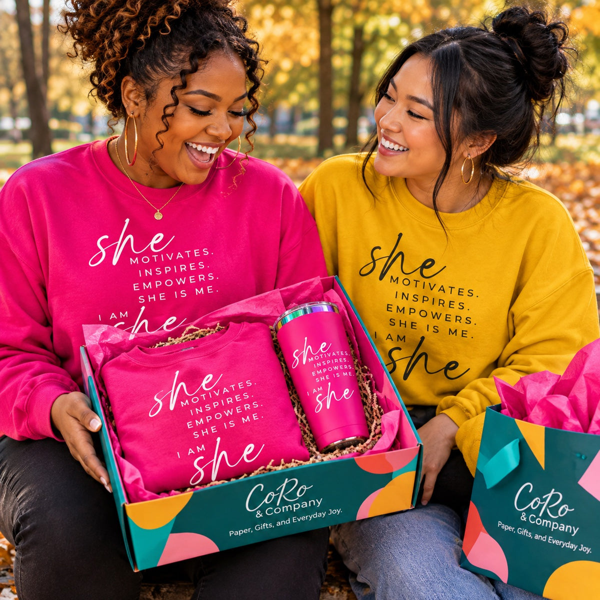 Two women sitting outdoors in a park setting, smiling and holding a CoRo & Company gift box. The box contains a matching hot water tumbler and a folded sweatshirt.