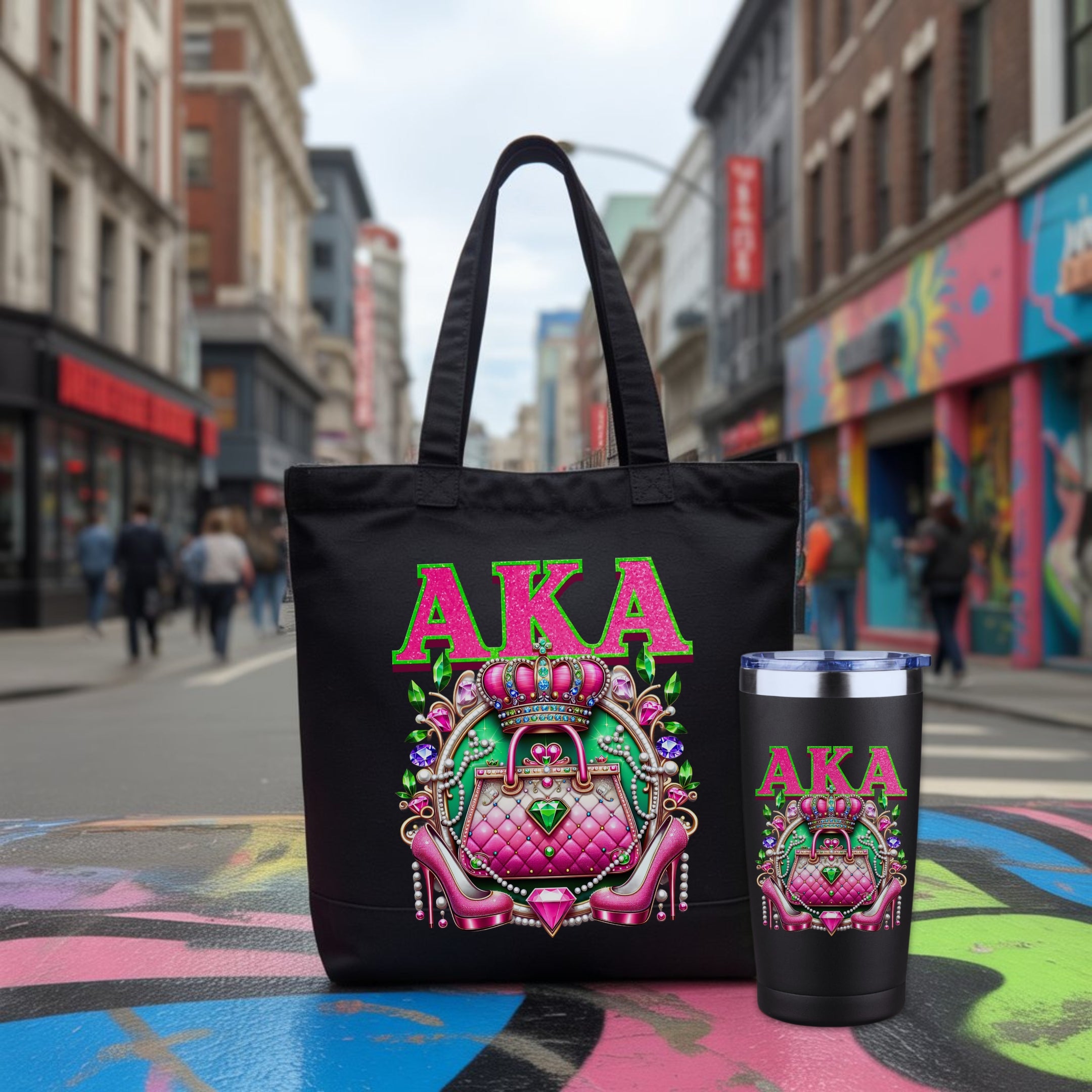 A black tote bag and matching black tumbler with a colorful pink and green graphic featuring the text 'AKA' and a purse illustration, photographed on a blurred city street with pedestrians and graffiti in the background.