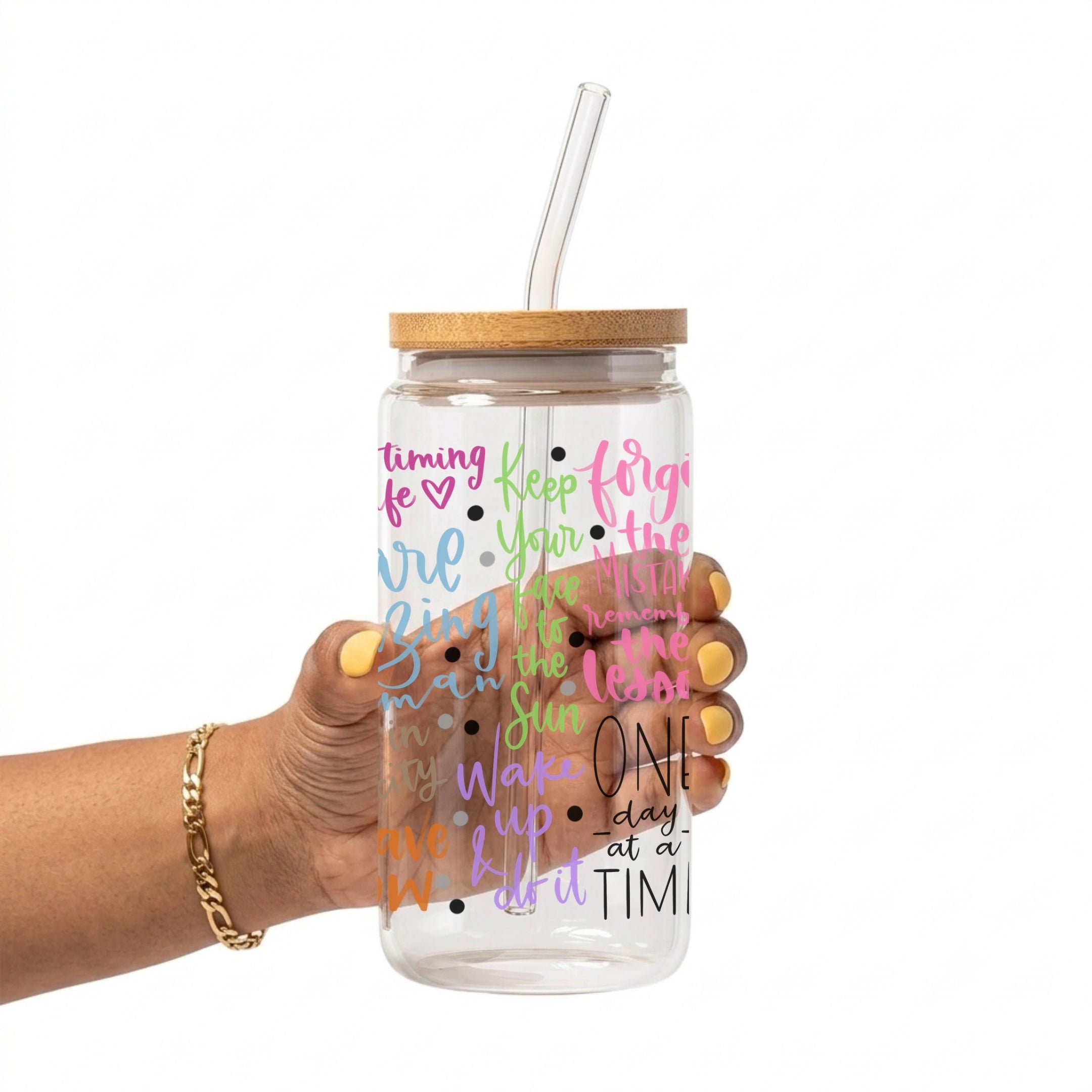 A hand holding a clear glass cup with a bamboo lid and clear straw. The tumbler features colorful motivational text printed on the front in pink, green, blue, and purple.