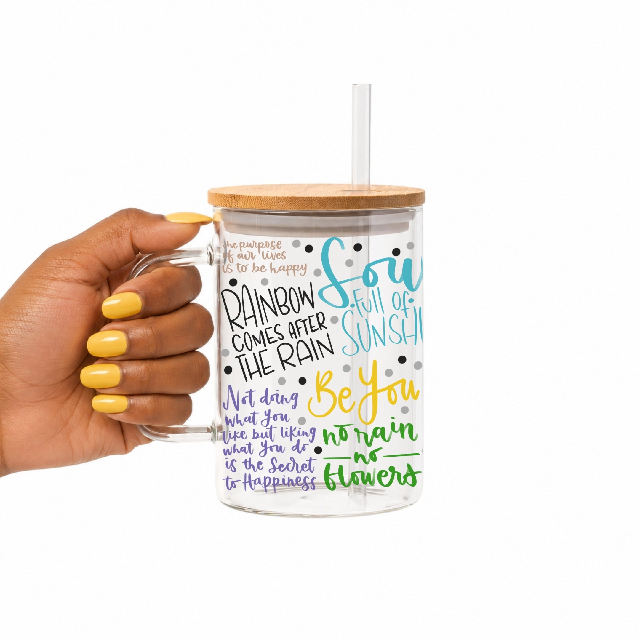 A hand with yellow nail polish holds a clear handled glass mug with a bamboo lid and clear straw. The tumbler is printed with colorful inspirational quotes in blue, green, yellow, and purple text with black polka dots on a white background.