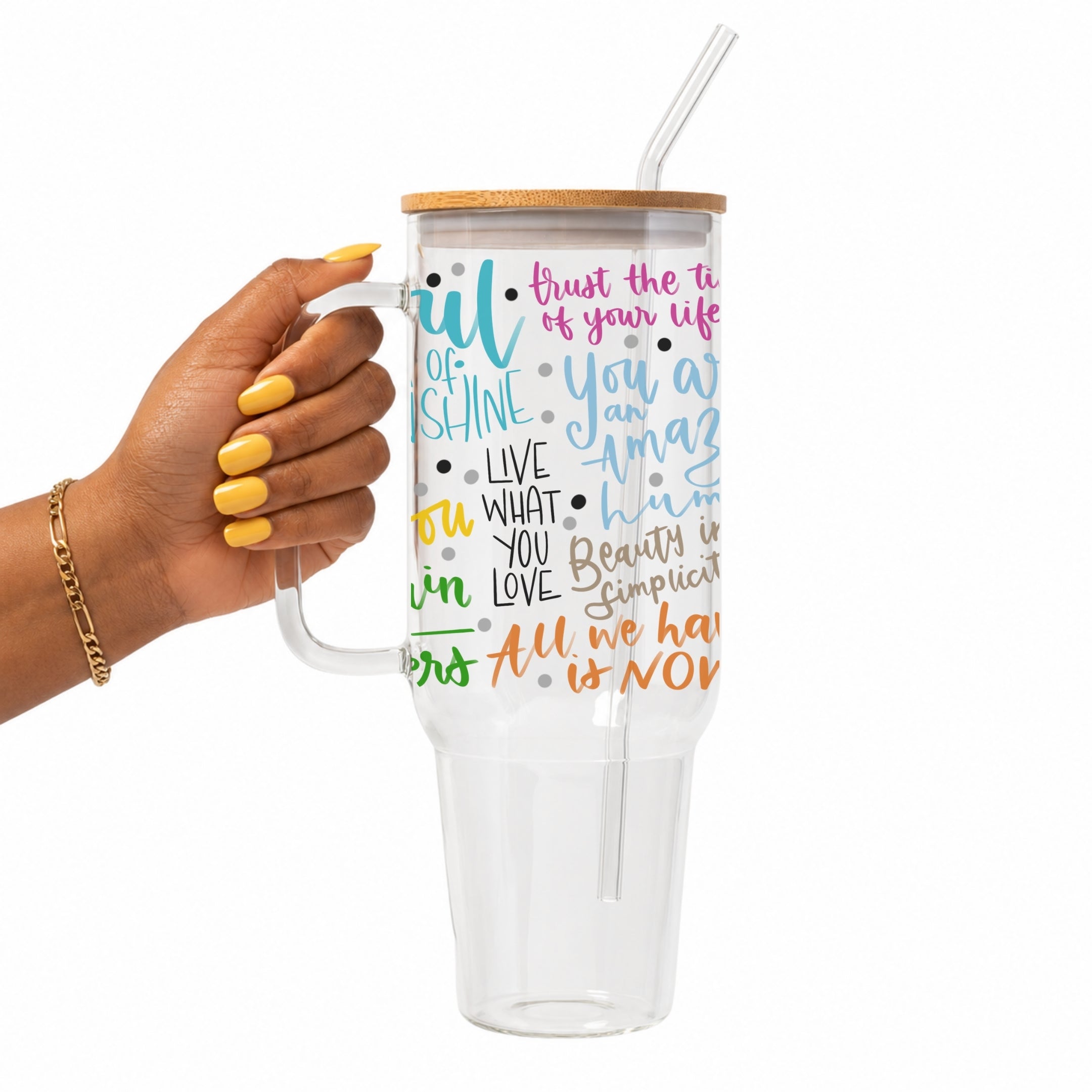 A hand with yellow nails holds a clear handled tumbler with a bamboo lid and clear straw. The tumbler is printed with colorful inspirational text on a white background.