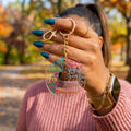 A person outdoors holds up a round clear acrylic keychain with colorful inspirational text. The keychain has a gold-tone split ring and chain.