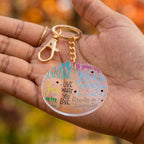 A round acrylic keychain with colorful inspirational text, held in a person's hand. The keychain has a gold-tone lobster clasp and chain, and the clear disc features multicolored script text on a warm-toned, blurred autumn background.