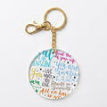 A round acrylic keychain with a gold-tone clasp and chain, featuring colorful hand-lettered motivational quotes on a white background.