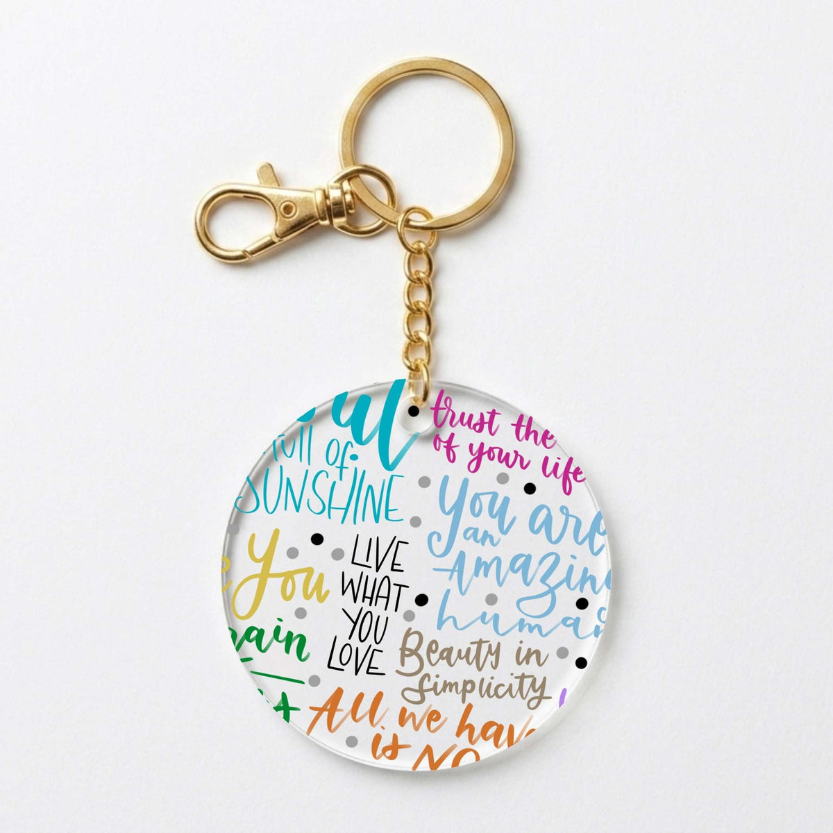 A round acrylic keychain with a gold-tone clasp and chain, featuring colorful hand-lettered motivational quotes on a white background.
