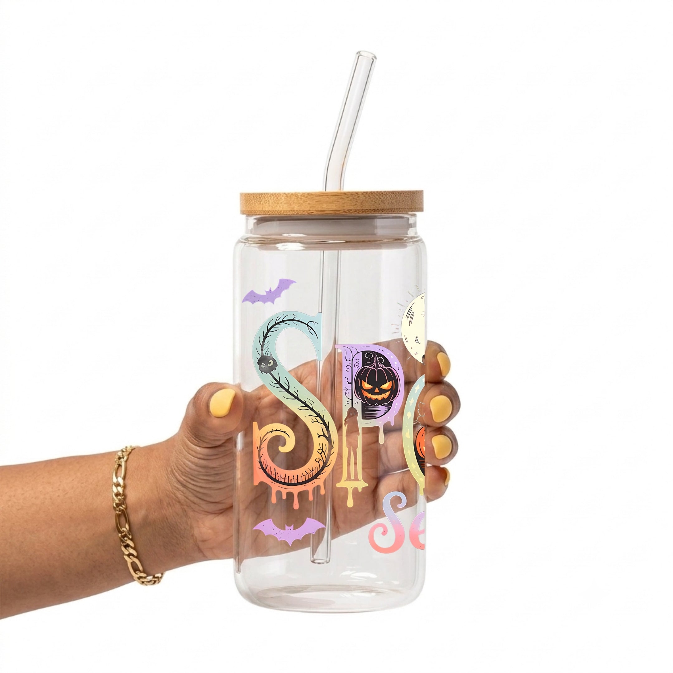 A hand holding a clear glass tumbler with a bamboo lid and clear glass straw, decorated with colorful Halloween-themed graphics including bats, a snake, a pumpkin, and the phrase 'Spooky Season'.