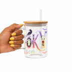 A hand with yellow nail polish holds a clear glass mug with a bamboo lid and clear straw. The mug features a colorful Halloween-themed graphic spelling 'SPOOKY' with a crow, purple lavender, a witch's hat, and the word 'Season' in pastel script.