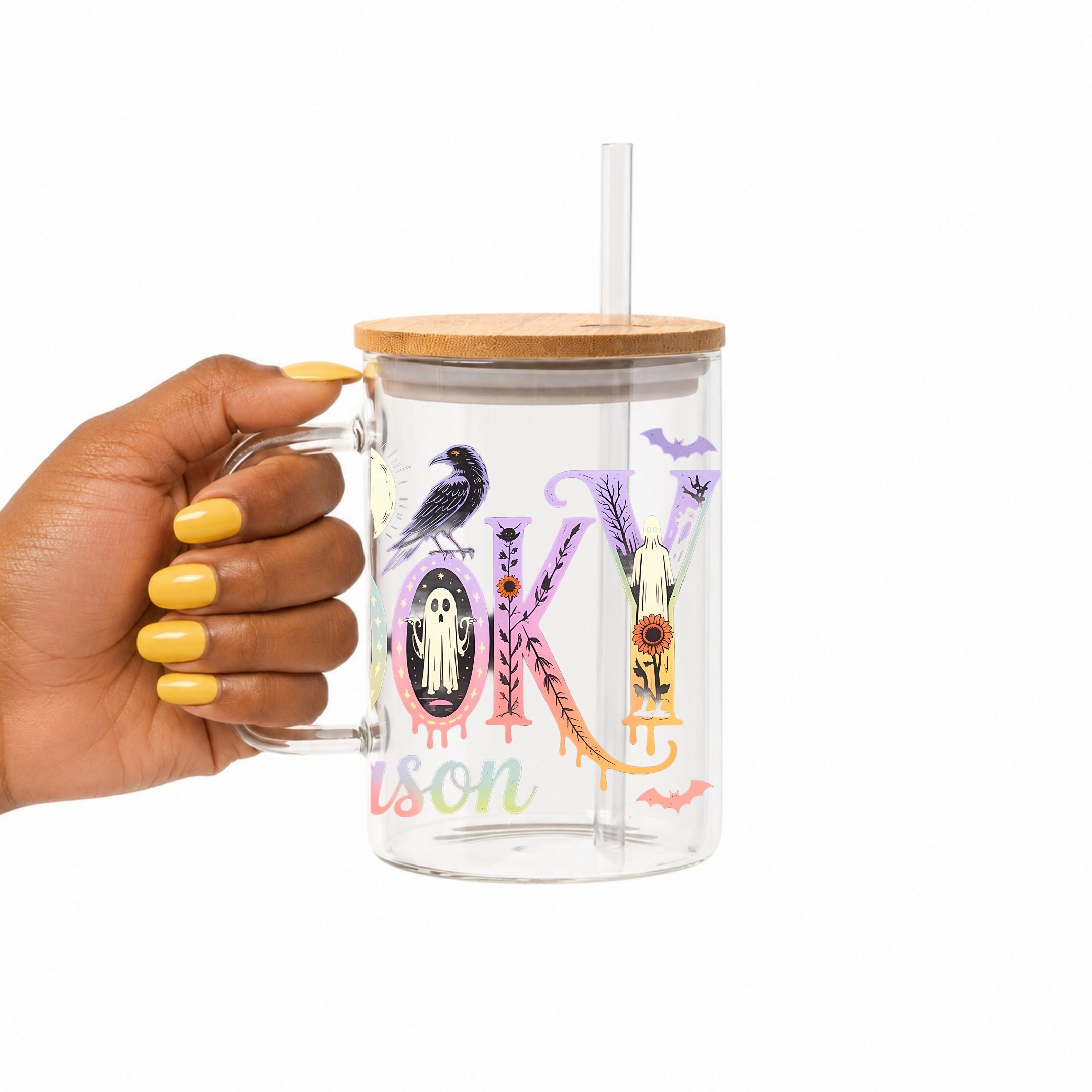 A hand with yellow nail polish holds a clear glass mug with a bamboo lid and clear straw. The mug features a colorful Halloween-themed graphic spelling 'SPOOKY' with a crow, purple lavender, a witch's hat, and the word 'Season' in pastel script.