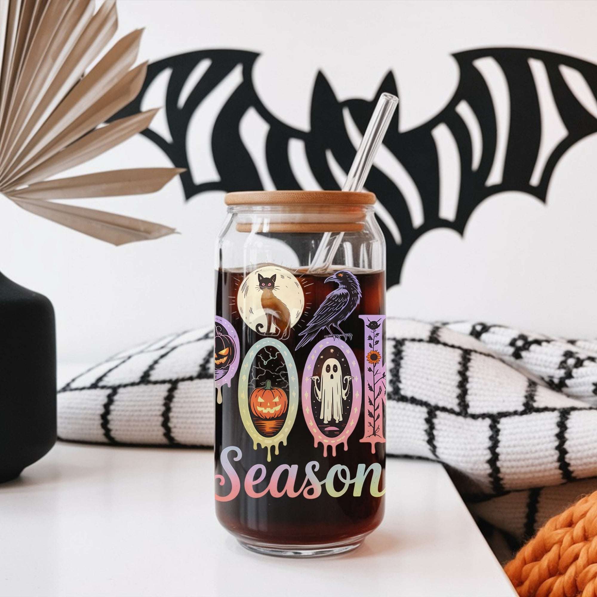 Glass tumbler with Halloween-themed design and straw, placed on a surface with decorative elements in the background.