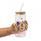 A hand holding a clear glass tumbler with a bamboo lid and clear straw, decorated with Halloween-themed graphics including a pumpkin, cat, spider, and the words 'Spooky Season' on a white background.