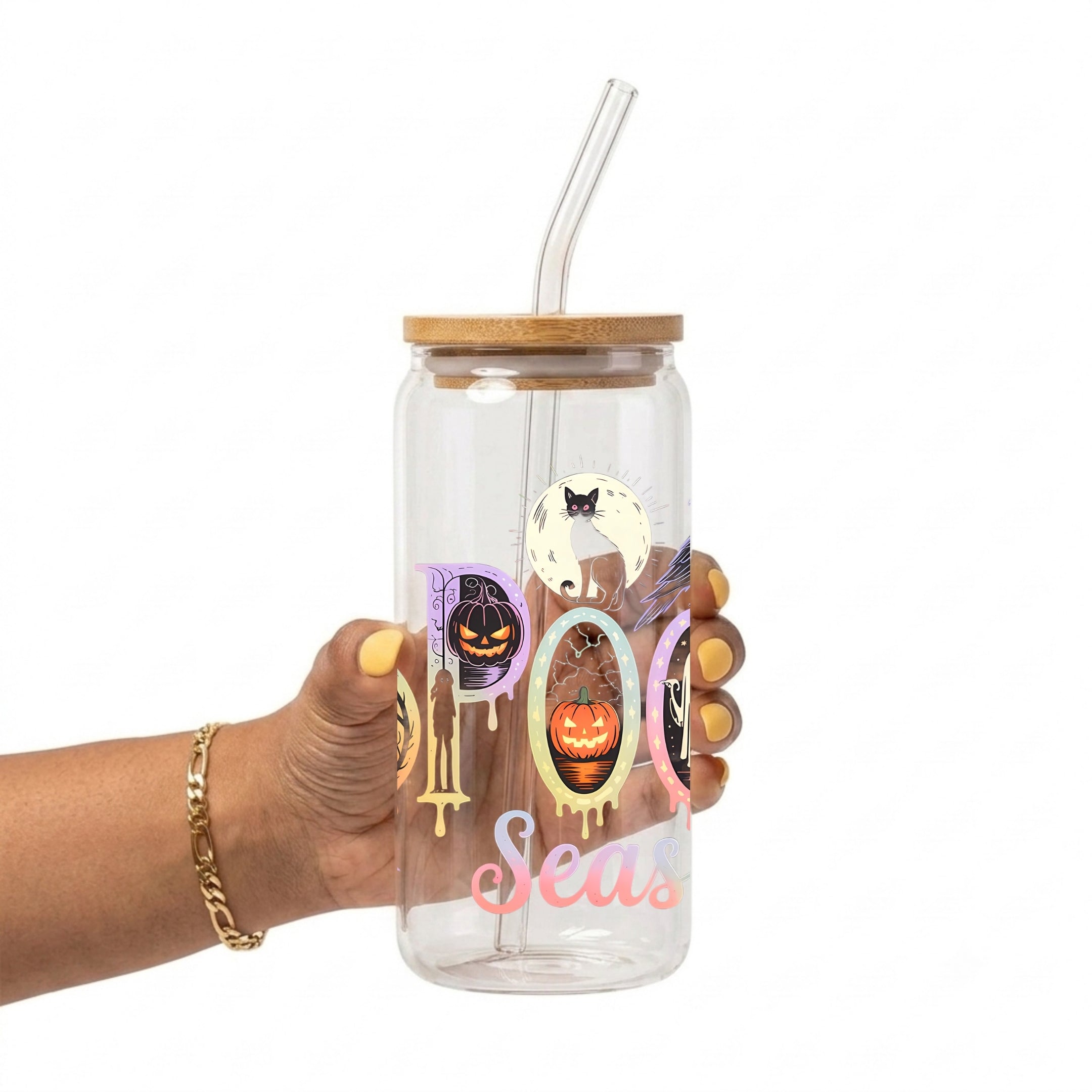 A hand holding a clear glass tumbler with a bamboo lid and clear straw, decorated with Halloween-themed graphics including a pumpkin, cat, spider, and the words 'Spooky Season' on a white background.
