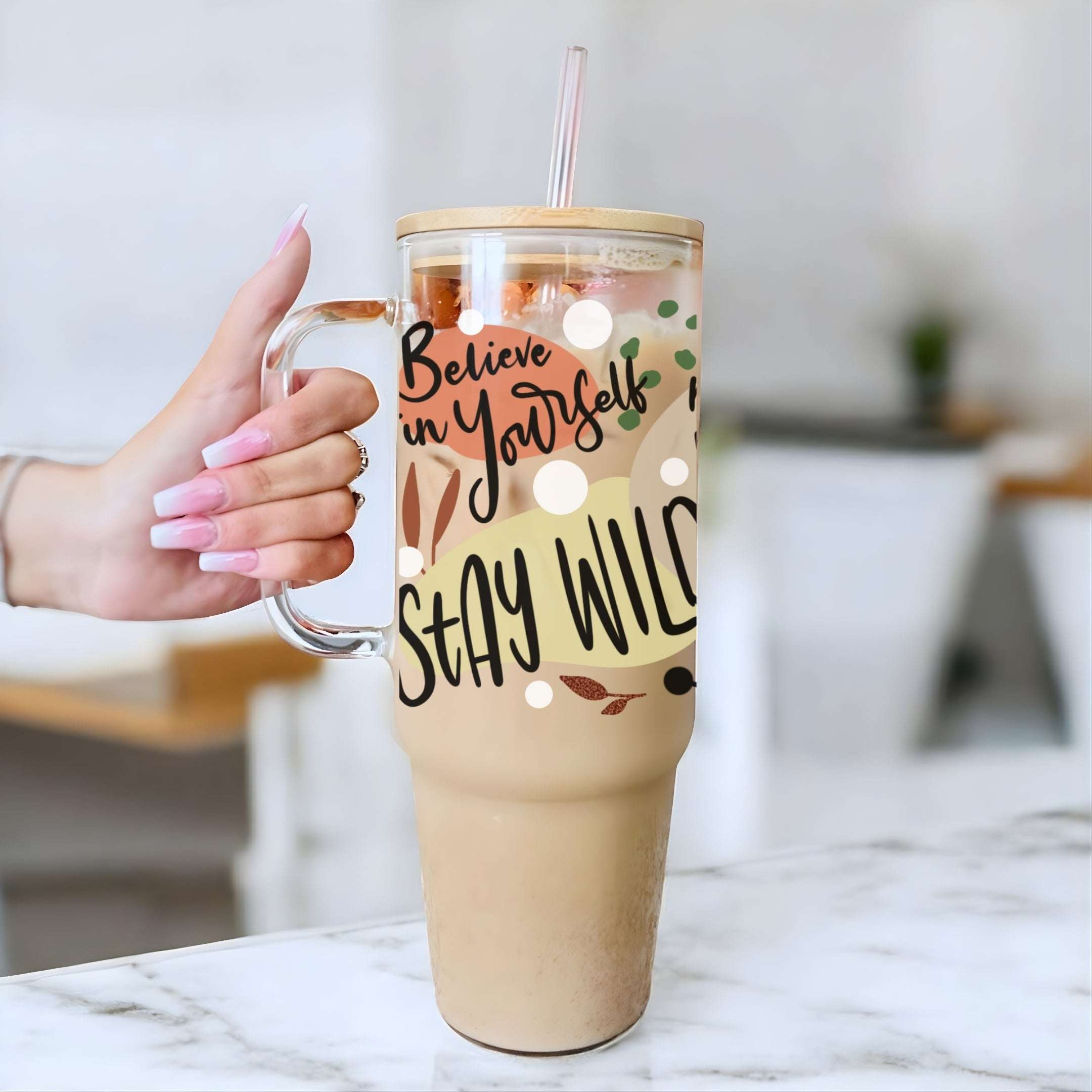 A hand with pink manicured nails holds a clear handled tumbler with a straw, filled with an iced coffee drink. The tumbler has a printed design with the words 'Believe in Yourself' and 'STAY WILD' in black script over a beige and brown leaf pattern.