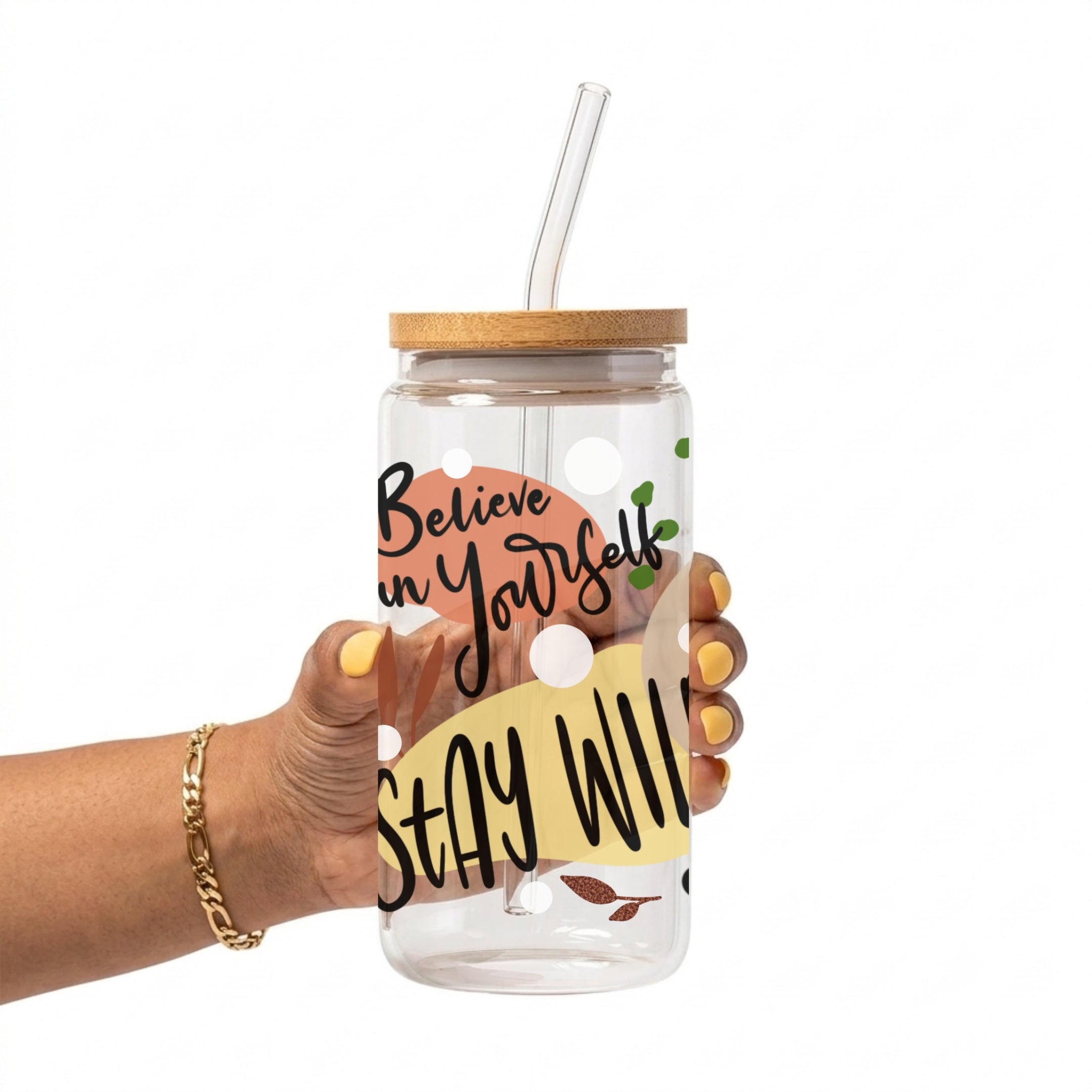 A hand holding a clear plastic tumbler with a bamboo lid and clear glass straw. The tumbler features a printed design with the text 'Believe in Yourself' and 'STAY WILD' on a yellow background with white polka dots.