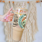 A hand with pink manicured nails holds a clear handled tumbler with a straw, filled with an iced coffee drink. The tumbler has a light wood lid, a clear glass handle, and a printed fall-themed graphic with the text 'TOMORROW IS A NEW DAY' and autumn leaves.