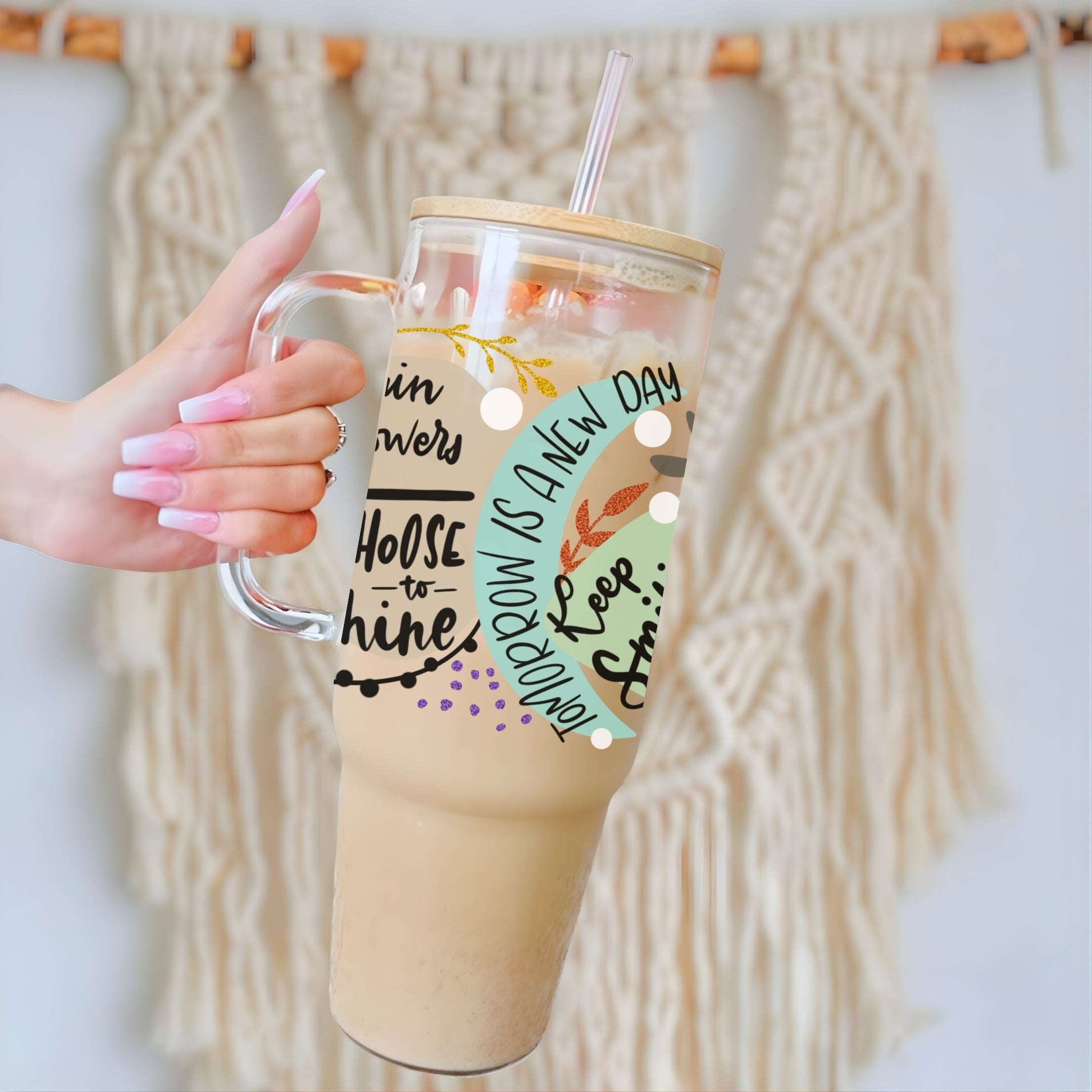 A hand with pink manicured nails holds a clear handled tumbler with a straw, filled with an iced coffee drink. The tumbler has a light wood lid, a clear glass handle, and a printed fall-themed graphic with the text 'TOMORROW IS A NEW DAY' and autumn leaves.