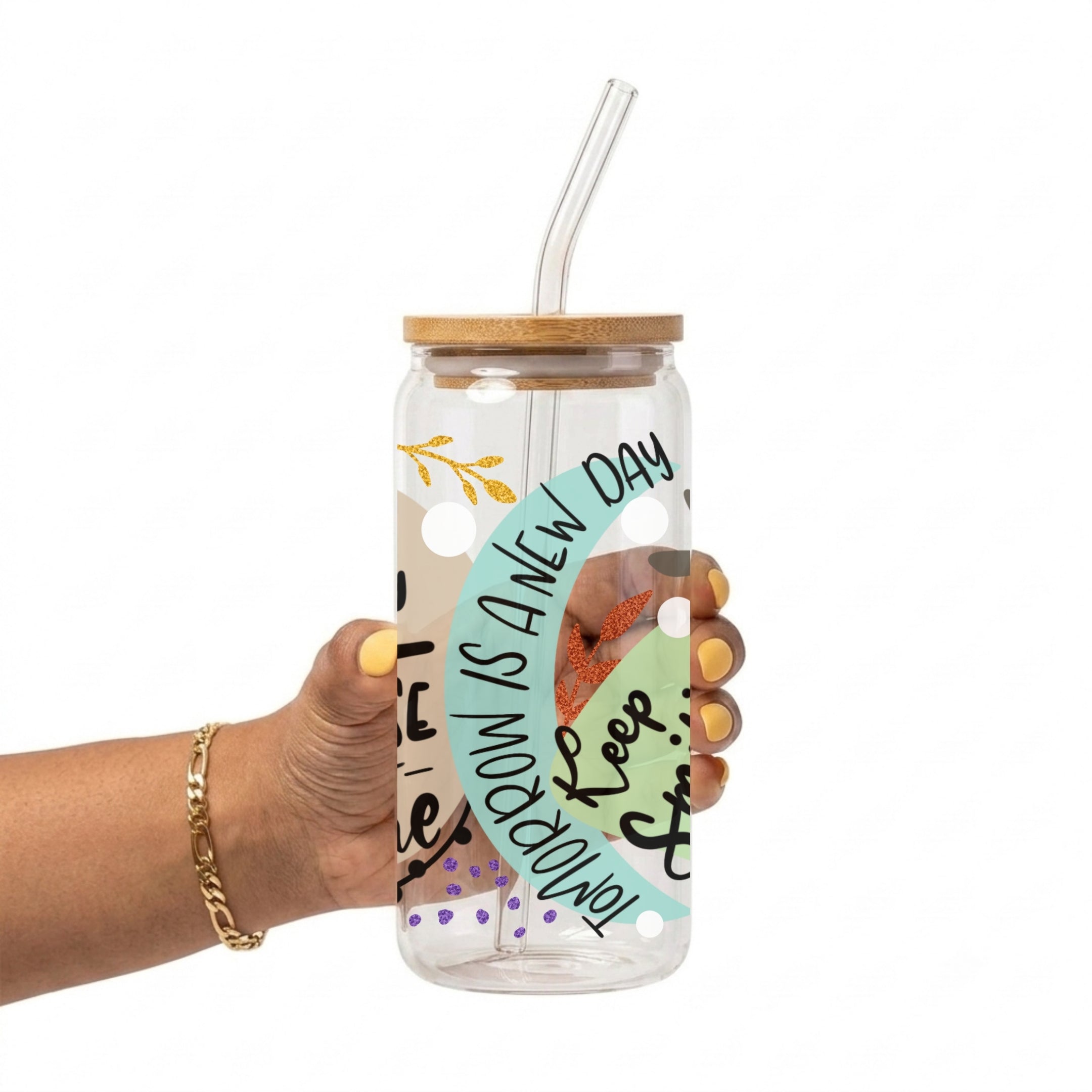 A hand holding a clear glass tumbler with a bamboo lid and clear straw. The tumbler features a colorful printed design with the text 'TOMORROW IS A NEW DAY' and 'Keep Smiling'.